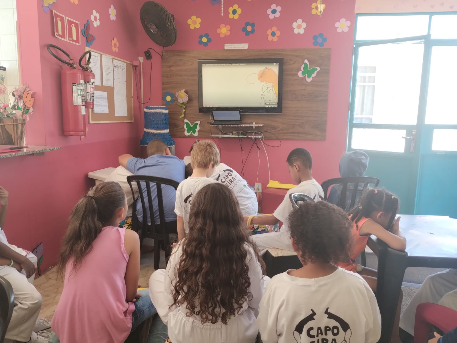 Na imagem, diversas crianças estão sentadas em uma sala de aula, com paredes azul e rosa, assistindo um vídeo em uma televisão